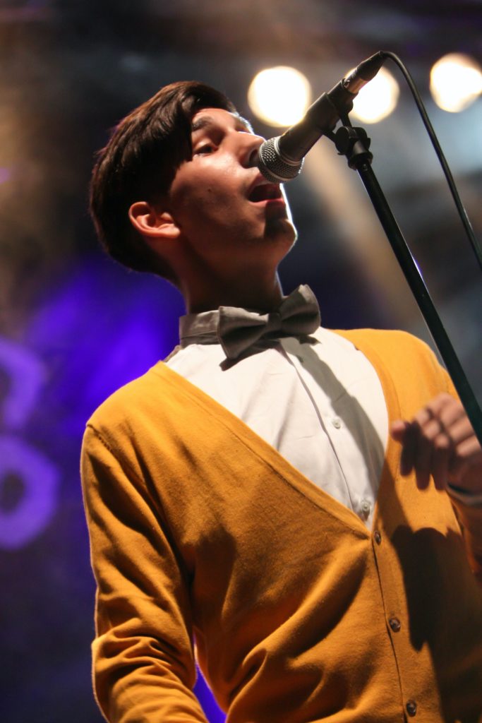 Man in yellow sweater and bowtie at a microphone.