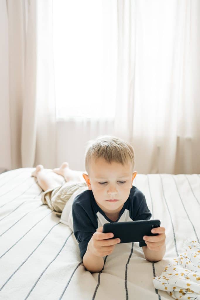Photo of a young boy laying on bed looking at a smartphone. 