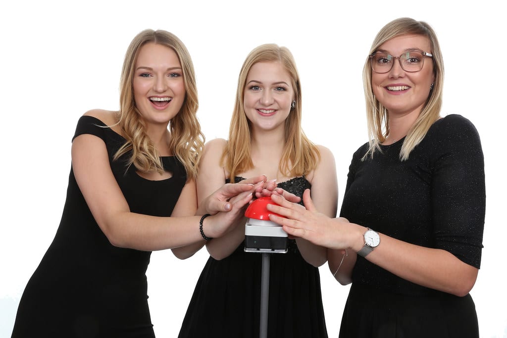 Three blonde women holding their hands over a red buzzer.