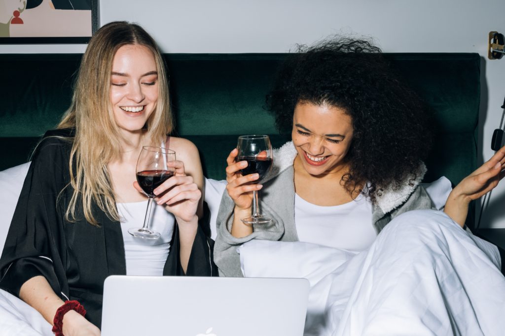 Two girls drinking wine and laughing in bed, looking at a laptop.