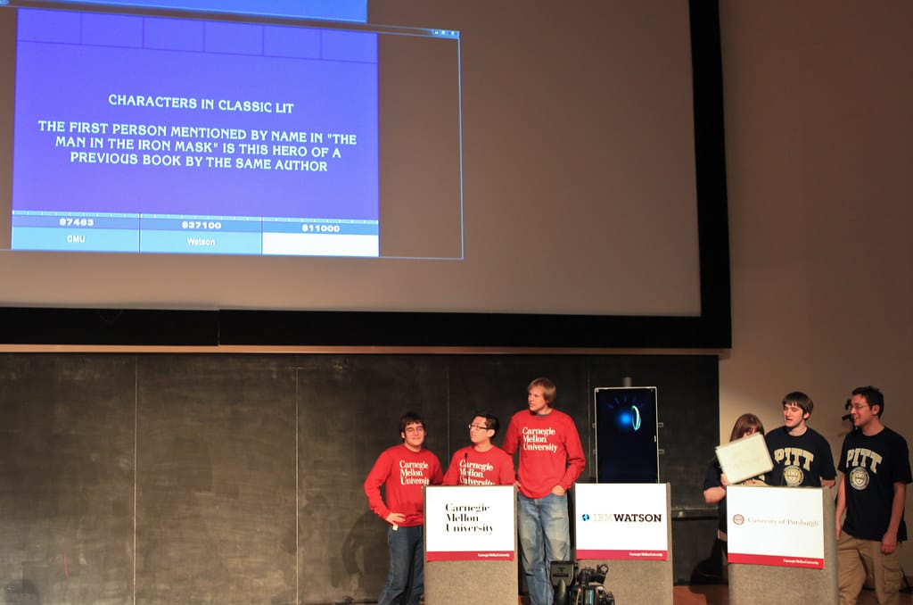 College students with Watson in front of a large screen displaying a Jeopardy question