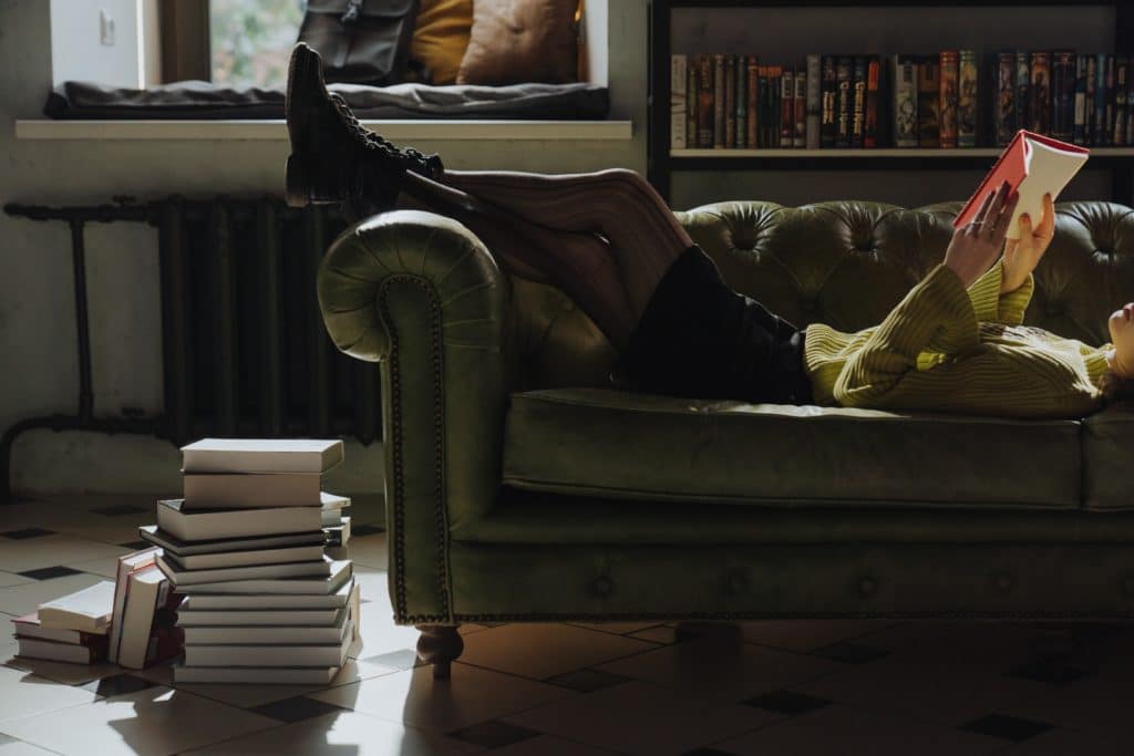 Woman lying on couch reading a book with a pile of books on the floor next to her