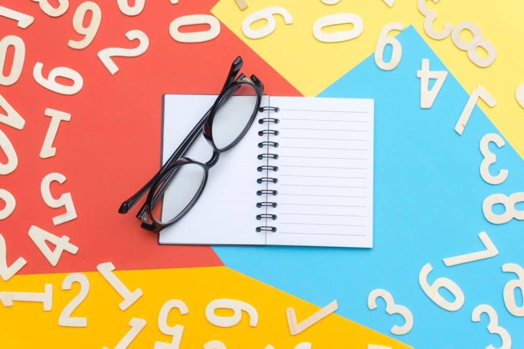 An open blank notebook with a pair of glasses resting on top of it on a blue, red, and yellow background with white numbers scattered around it.