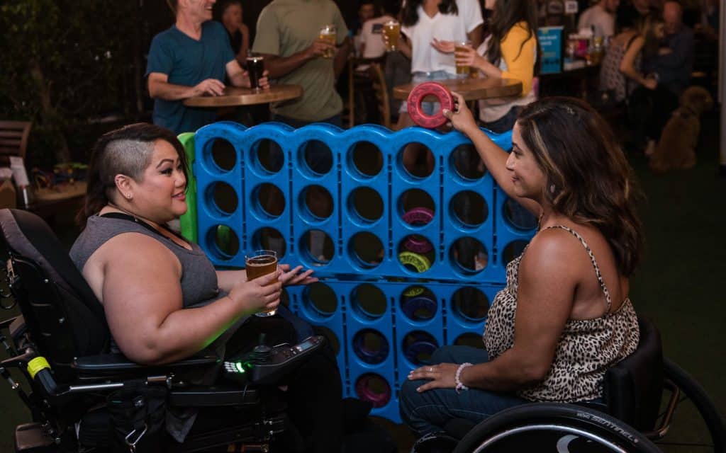 Two women in wheelchairs holding beers and playing a giant game of Connect 4.