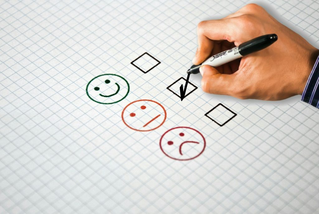A hand selecting a multiple choice answer of three options with a smiley face, an indifferent face, and a sad face.