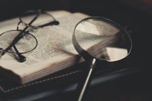 A pair of eyeglasses sitting atop an open book with a magnifying glass resting against it