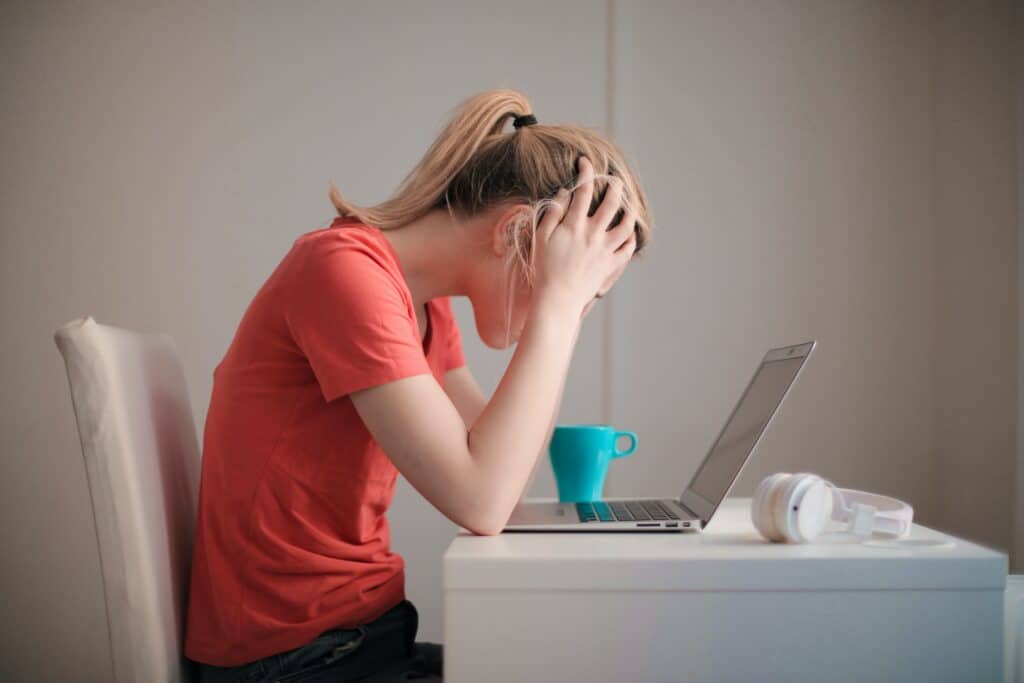 Blonde woman holds head while looking at a laptop, a teal coffee mug next to her. 