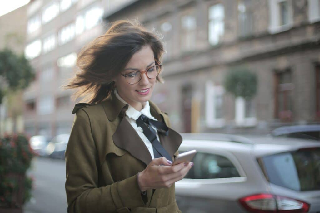 Woman walking wearing glasses, looking at her smart phone and smiling