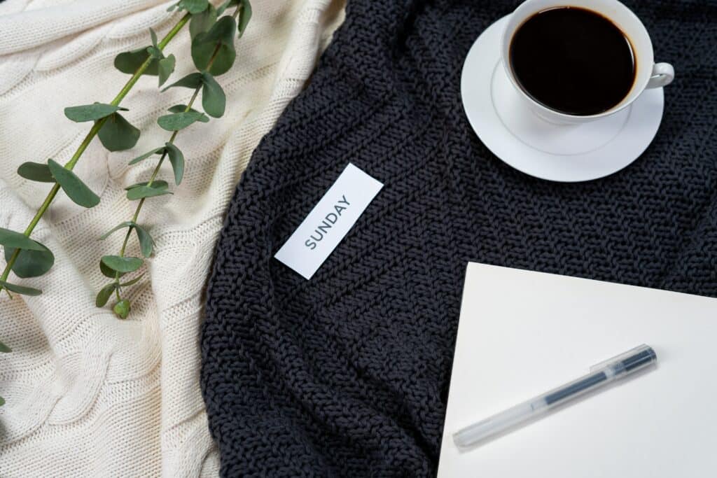 A slip of paper that reads "SUNDAY" atop knit blankets along with eucalyptus leaves, a cup of coffee and a blank piece of paper with a pen on top of it.