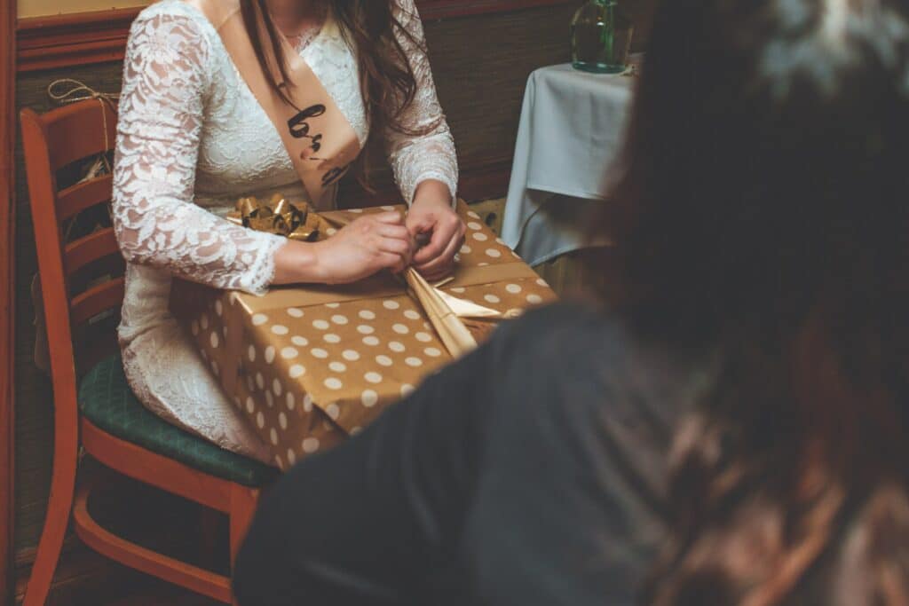 A bride-to-be in  white lace dress wearing a sash that says "bride" and opening a gift
