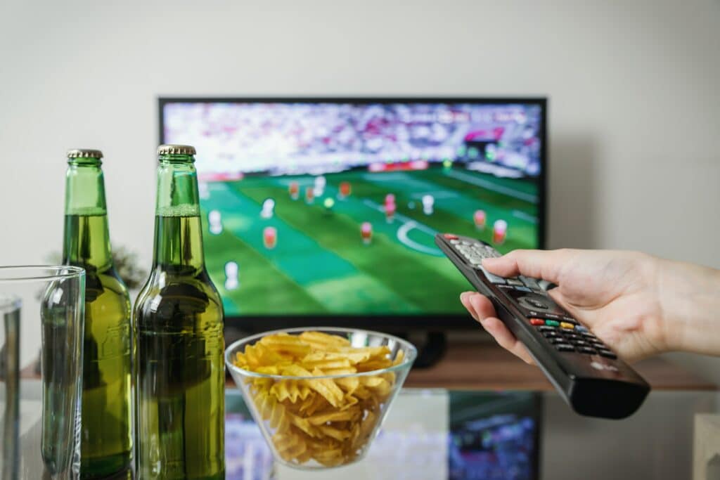 A person holding a remote up to a TV playing soccer. Chips and two bottles of beer are on a table in front. 