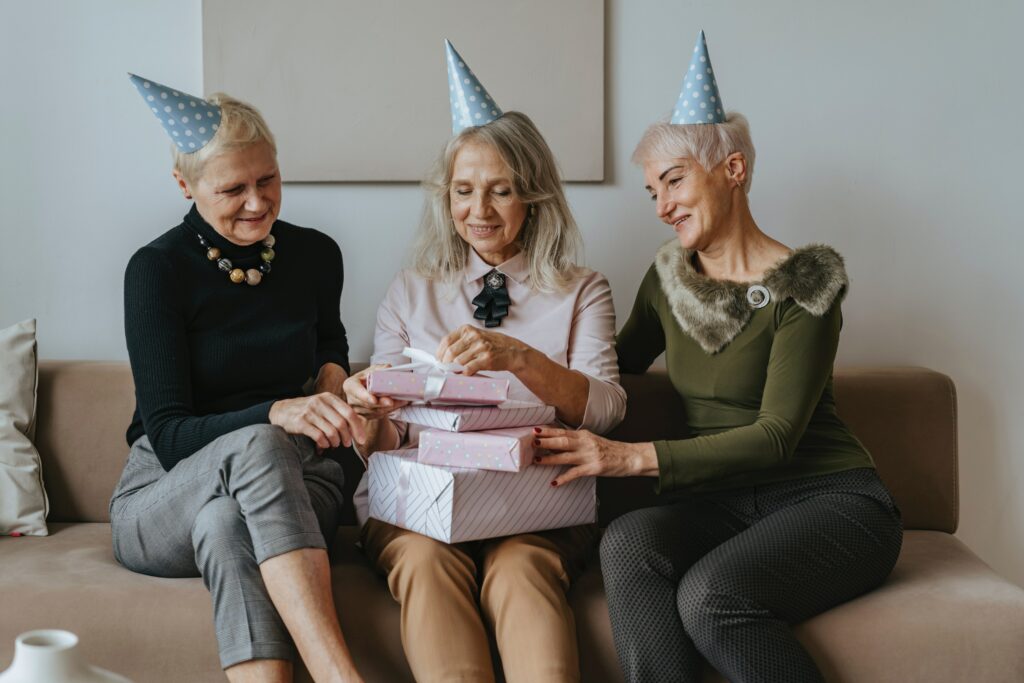 Three older women wearing birthday hats, the woman in the middle is opening presents