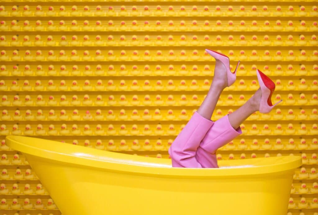 A woman's legs in pink pants and pink heels coming out of a yellow tub in front of a wall of yellow rubber duckies