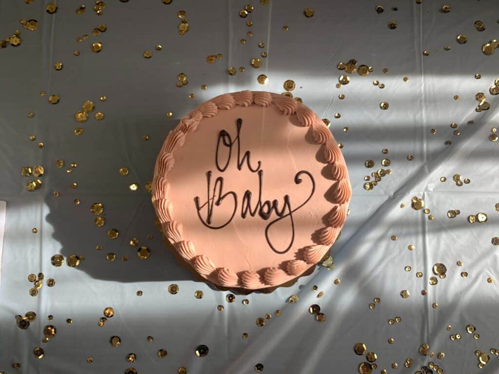Pink mousse cake with the words "Oh Baby" written in chocolate, atop a white tablecloth with gold confetti