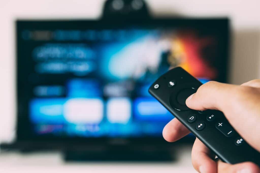 Close up of a hand holding a streaming device remote, pointed at an out-of-focus large-screen TV in the background