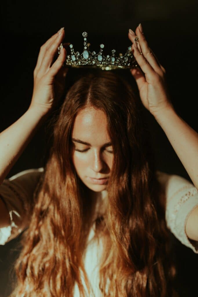 Photo of a young woman with long brown hair placing a crown on her head. 