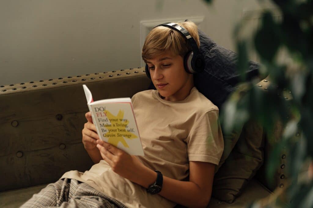 Photo of a blond teenage boy sitting on a couch reading a book with headphones on.