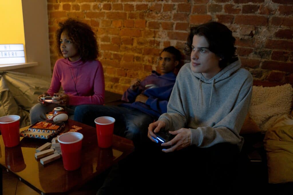 Photo of three teens sitting on a couch in front of a brick wall, playing video games. 