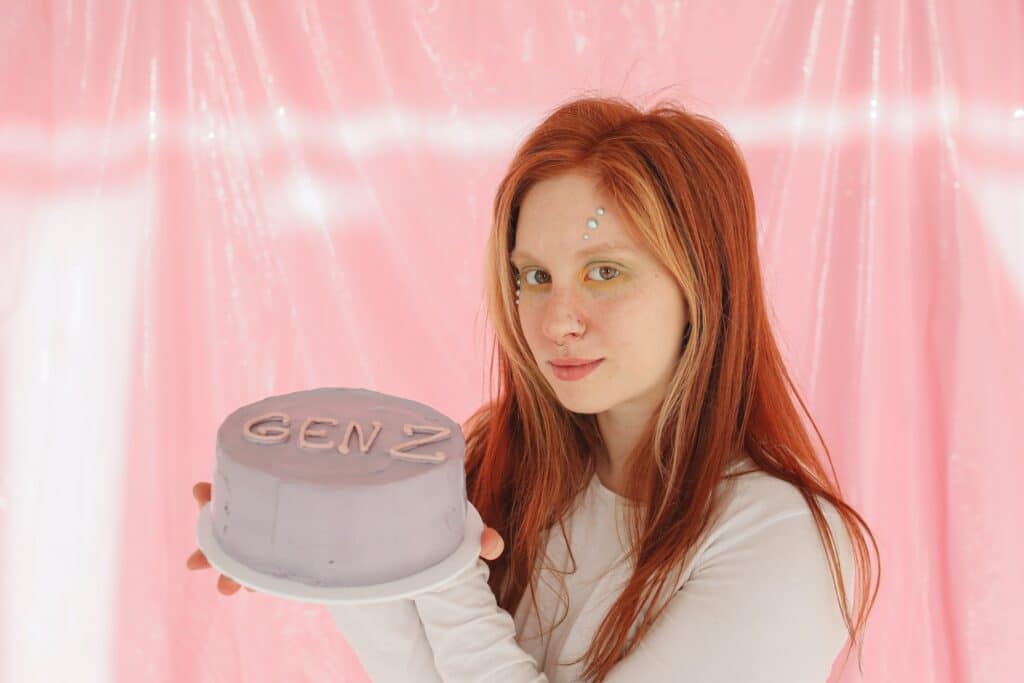 A young woman with red hair is holding a purple cake that reads "GEN Z" in pink icing. She's standing in front of a pink curtain. 
