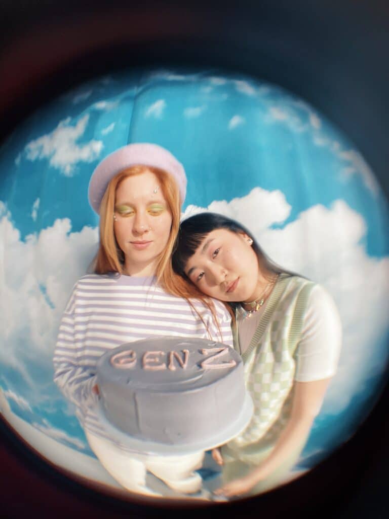 A fisheye-style photo of two young women holding a purple cake that says "GEN Z" in pink icing, against a sky-pattern bakdrop. 