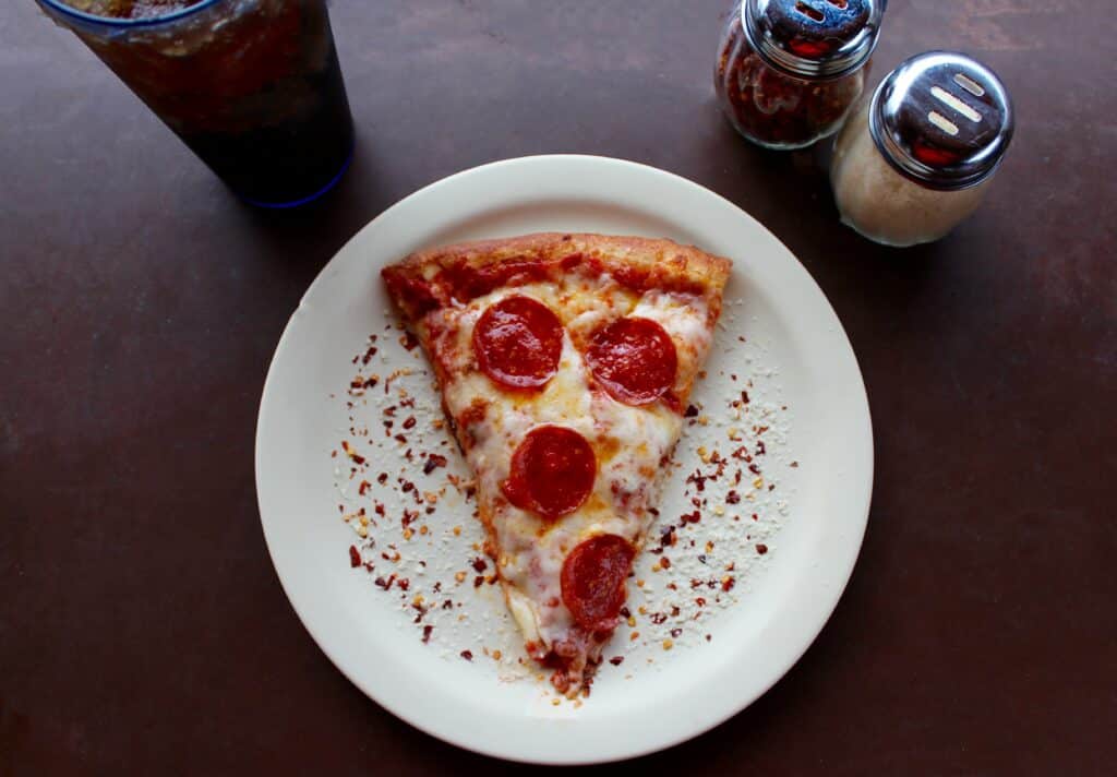 Photo of a slice of pepperoni pizza on a white, surrounded by soda and pizza seasonings.