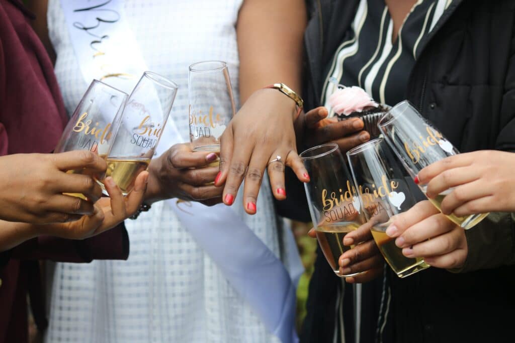 Photo of a woman in a white dress showing off her wedding ring, along with other women holding champagne glasses that read "bride squad"
