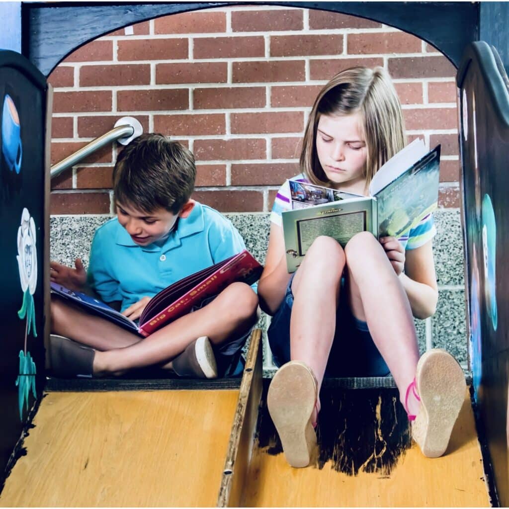 Photo of two kids sitting at the top of  slide, reading books. 