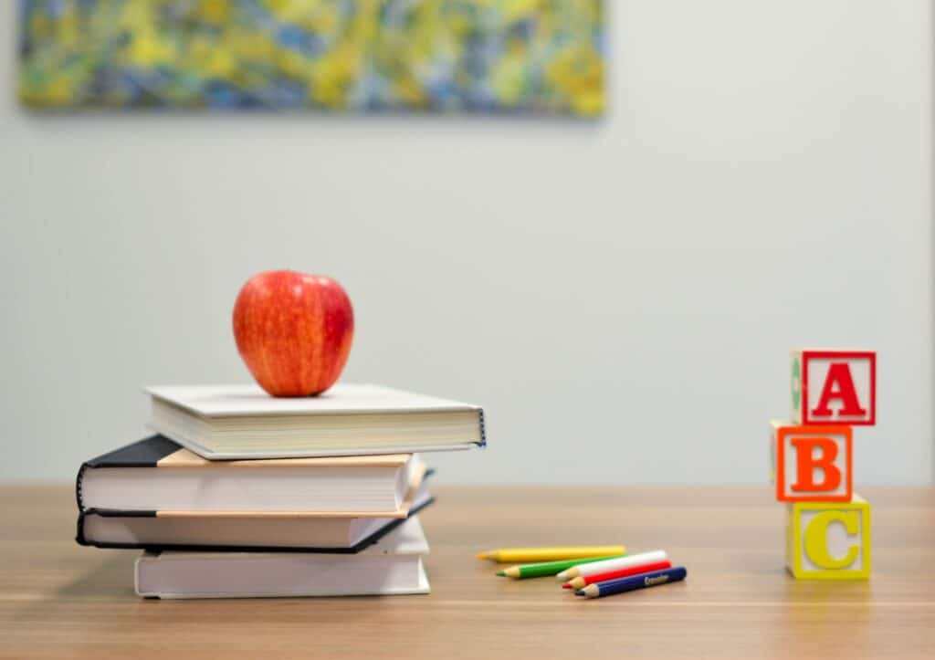 An apple on top of books on a desk, next to colored pencils and spelling blocks