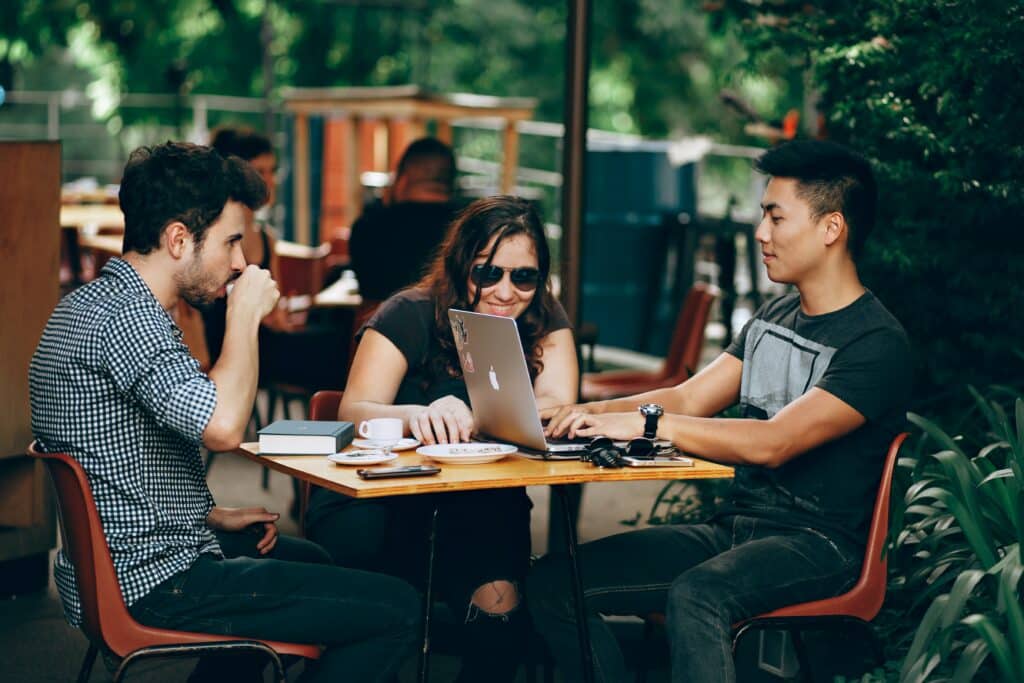Photo of three Millennial-aged people, two guys and a girl, sitting at an outdoor coffee shop looking at a laptop