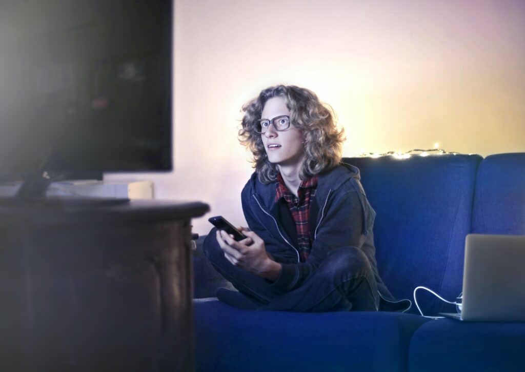 Person with blonde hair and glasses sitting on a blue couch next to a laptop, holding a remote and watching TV.