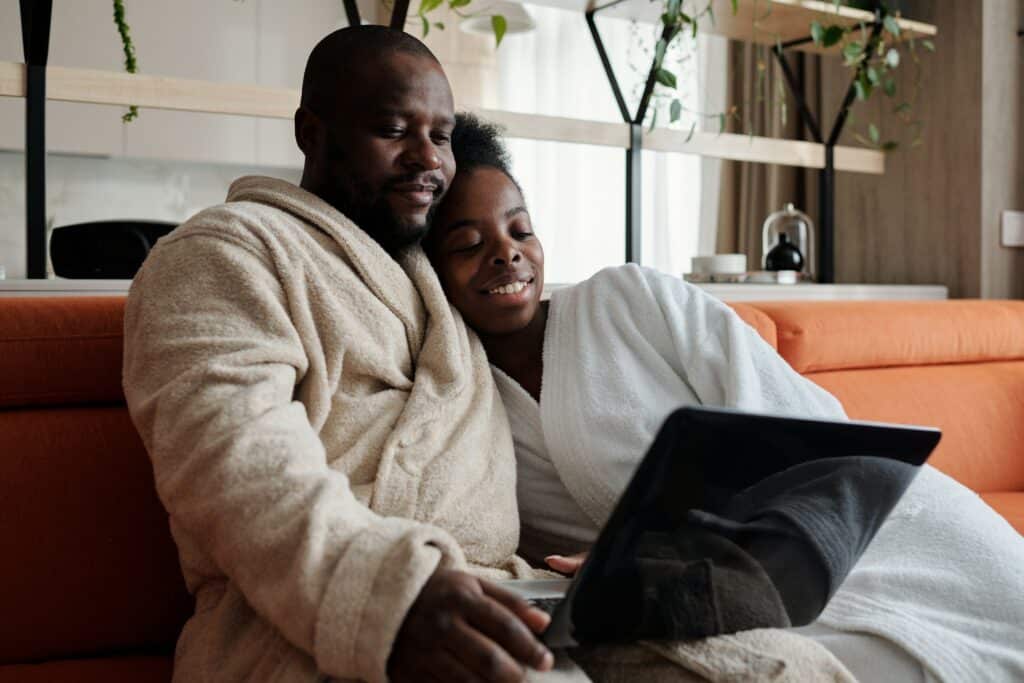 Photo of a couple in bathrobes snuggling up on an orange couch, watching something on a laptop.