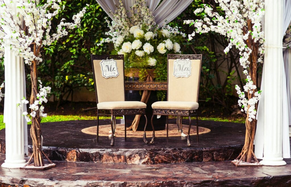 Photo of two chairs at an outdoor wedding, once with a sign on it that says Mr. and another with a sign on it that says Mrs.