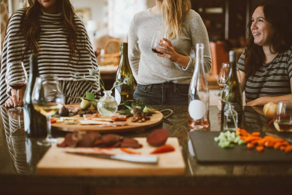 Photo of three women drinking wine and talking in a kitchen, surrounded by food and wine bottles