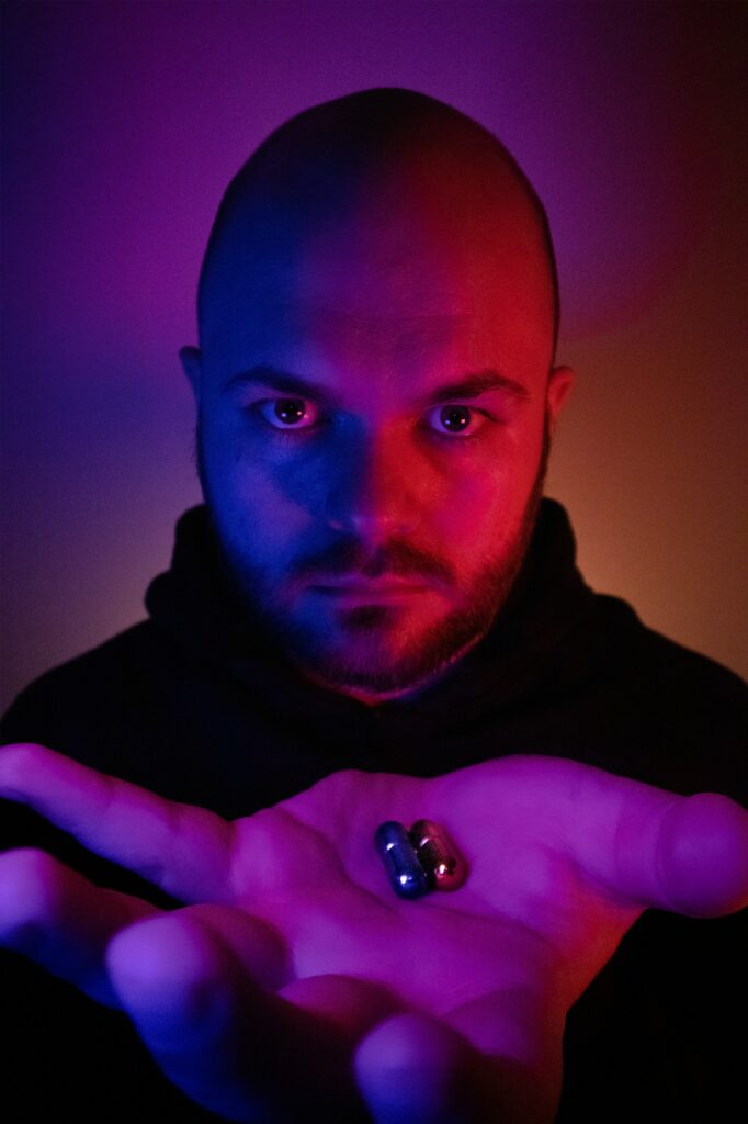 Neon-backlit photo of a man holding his hand out, with a red pill and blue pill in his palm. 