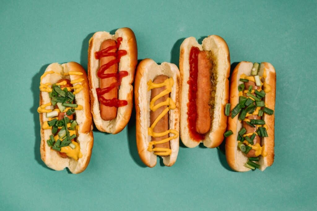 Five hot dogs with various toppings in a row against a teal background