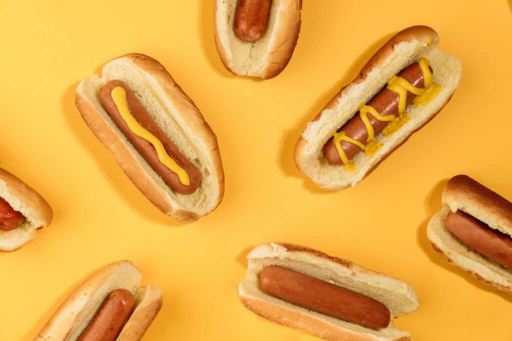 Photo of multiple hot dogs on buns, some with mustard, against a yellow background.