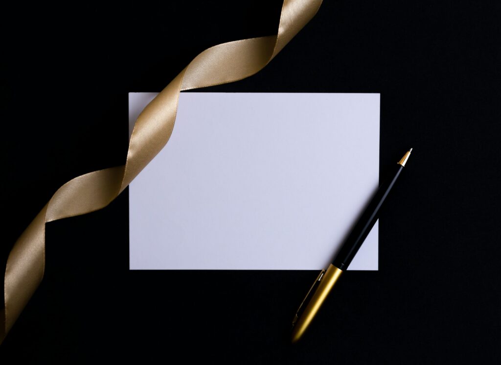 Photo of a blank white envelop with a pen and a gold ribbon atop it, all against a black background.