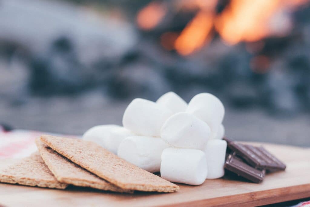 Photo of graham crackers, marshmallows, and chocolate bars on a wooden slab in front of a campfire