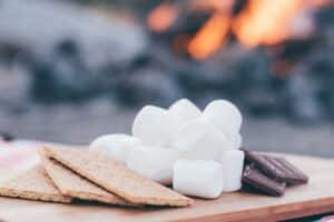 Photo of graham crackers, marshmallows, and chocolate bars on a wooden slab in front of a campfire