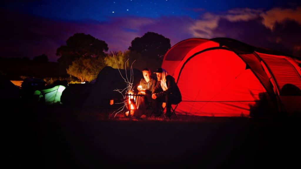 Photo of a couple sitting in camp chairs beside a campfire in front of a red tent with the night sky behind them.