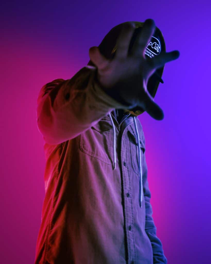 Man in a jacket with his hood up over his hat, obscuring his face with his hand against a pink and blue gradient background