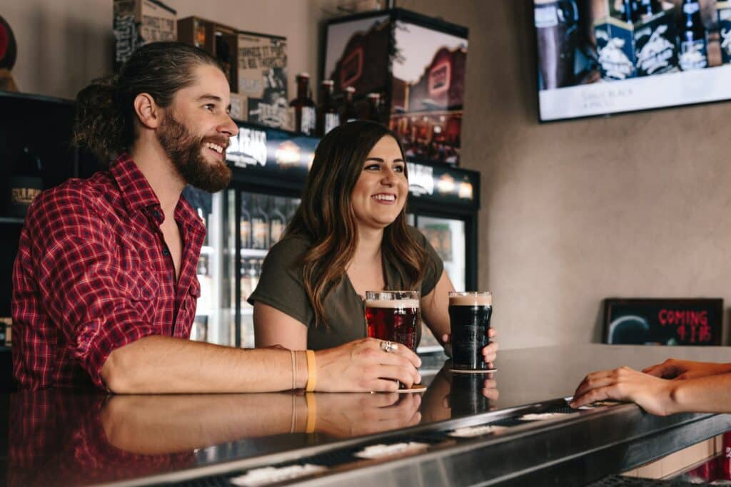 A woman and man sitting together at a bar drinking beers.
