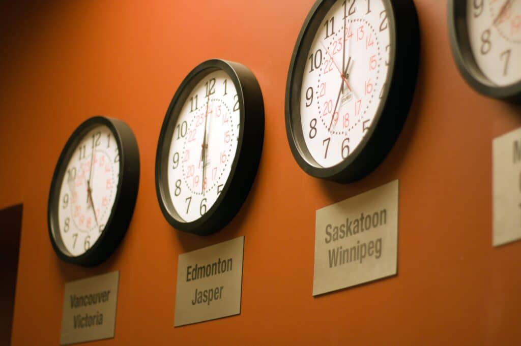 Round analog clocks on an orange wall displaying times in Vancouver, Victoria, Edmonton, Jasper, Saskatoon, Winnipeg. 