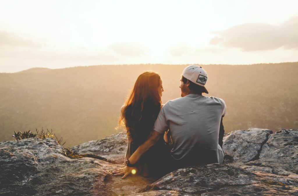 Photo of a young couple sitting next to each other on rocks at the top of a mountain at day.