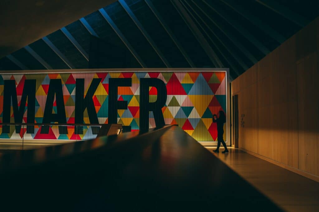 Photo of a person in a gallery walking past a large sign that reads "MAKER" in black text with various multicolored triangles behind it. 