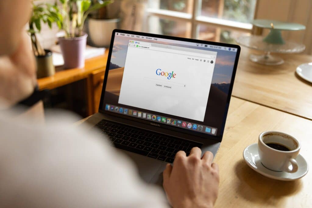 Photo of a person sitting at a desk with a cup of coffee next to an open laptop displaying a Google search page. 