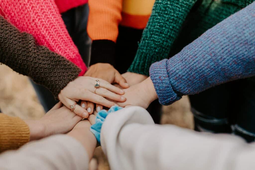 A group of people of diverse backgrounds wearing colorful sweaters putting their hands together.