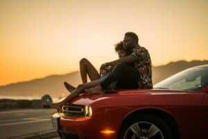 Photo of a couple in an embrace, sitting atop the hood of a red car with the sun setting in the background.