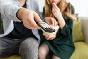 A man and a woman sitting on the couch, the woman holding a bowl of popcorn, the man pointing the remote at a TV behind the scenes