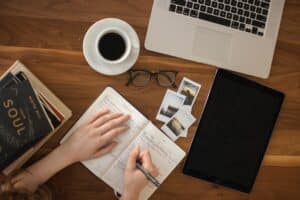 photo of a woman's hands writing in a notebook on a desk. On the desk is an open laptop, a cup of black coffee, a pair of glasses, photos, an iPad, and some self-help books.